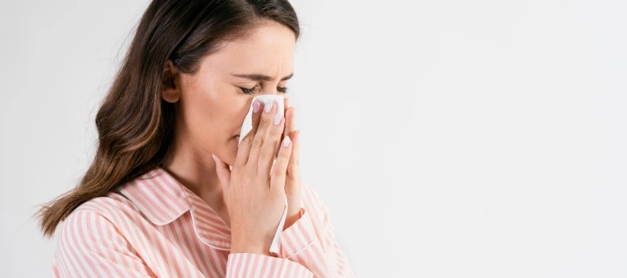 Mulher assoa o nariz com lenço de papel, usando pijama listrado, fundo claro. 