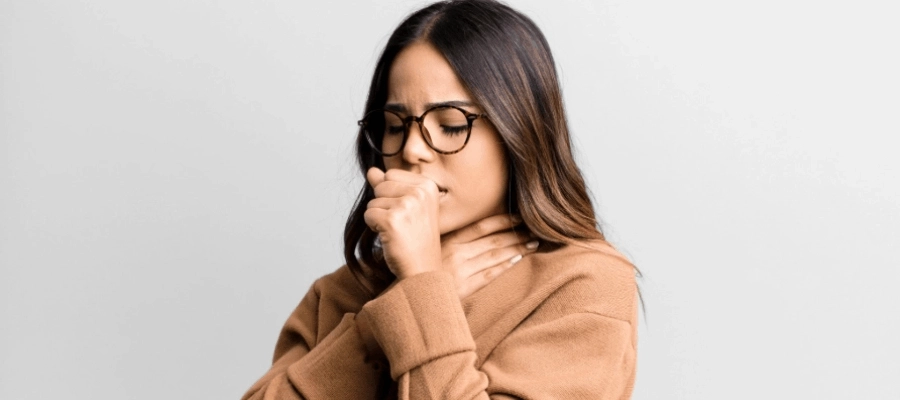 Mulher com óculos tosse e segura a garganta, vestindo blusa marrom, fundo claro.