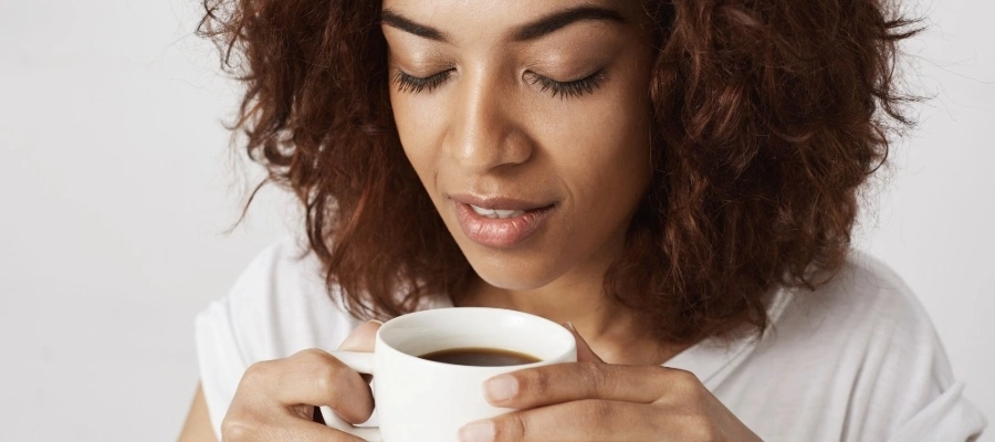 Mulher de cabelos cacheados segura xícara de café, com olhos fechados e expressão de contentamento. 
