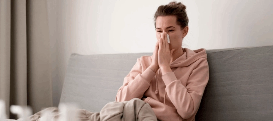 Mulher está sentada em um sofá enquanto assoa o nariz entupido, sintoma clássico da gripe.