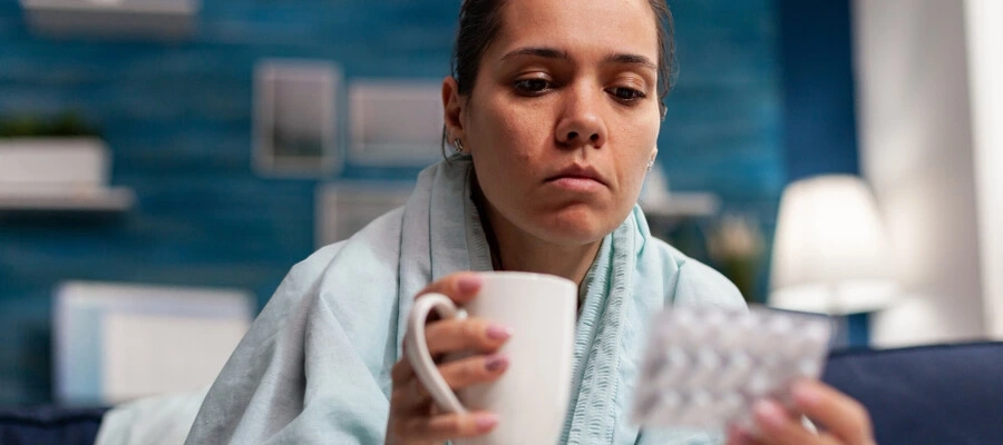 mulher gripada em casa tomando chá e segurando uma cartela de remédio