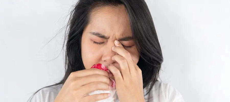 Mulher com expressão de dor pressionando o nariz e segurando um pano com sangue, indicando sangramento nasal.