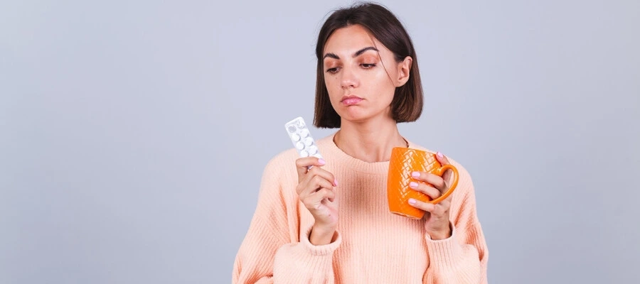 Mulher com cabelo curto segurando xícara e comprimidos com expressão de dúvida