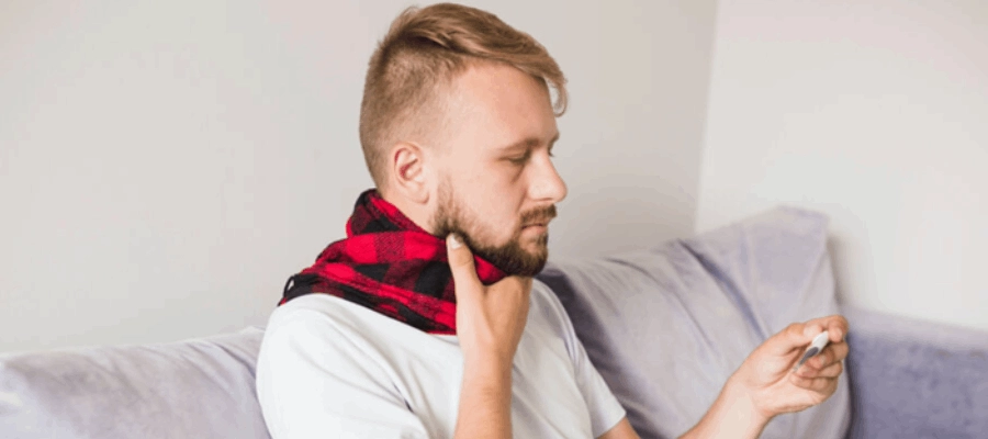 homem com febre e garganta inflamada checando a temperatura com um termômetro.