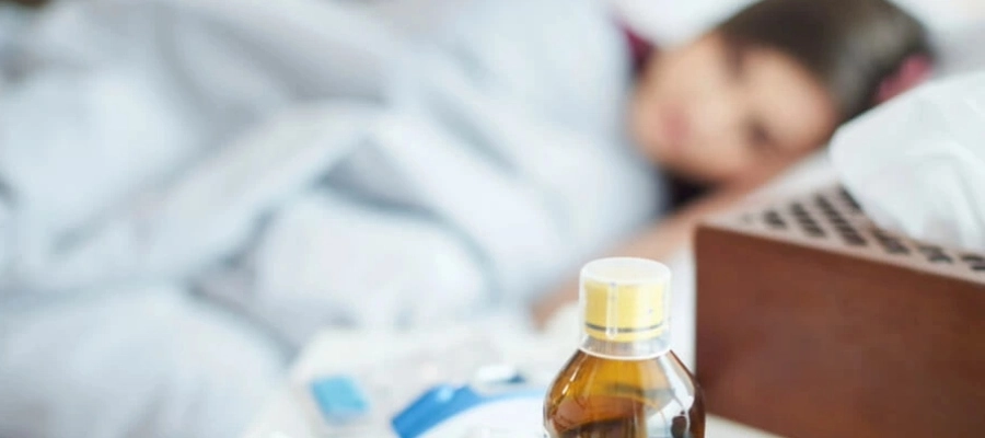 mesa com xarope para gripe e remédios, com mulher deitada na cama ao fundo