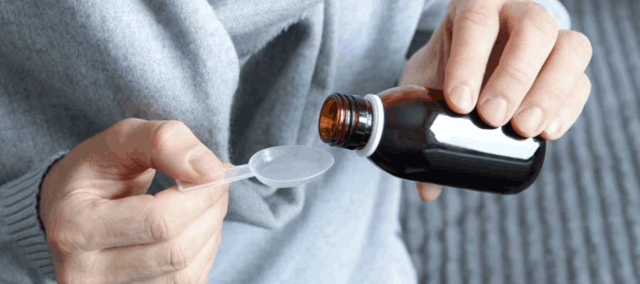 Mãos de uma pessoa que despeja xarope de frasco âmbar em uma colher dosadora plástica.