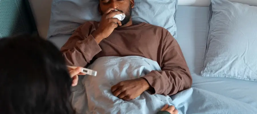 Homem deitado na cama com expressão de mal-estar, cobrindo-se com cobertor e segurando lenço no rosto, enquanto outra pessoa segura um termômetro.