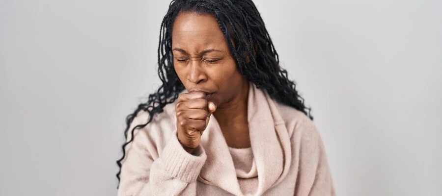mulher com cabelos compridos vestindo uma blusa bege com a mão em frente a boca enquanto tosse