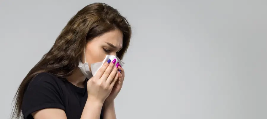  Mulher com expressão de incômodo usando máscara e assoando o nariz com lenço, indicando sintomas de alergia respiratória