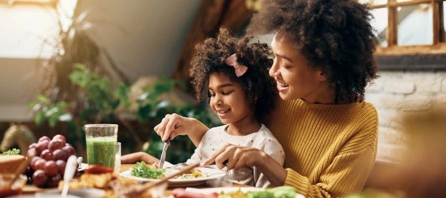 Mãe e filha sorriem enquanto fazem refeição juntas em uma mesa com alimentos saudáveis. 
