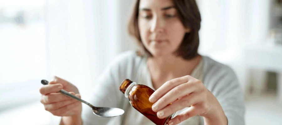 Mulher despeja xarope de uma garrafa em uma colher, em ambiente iluminado.