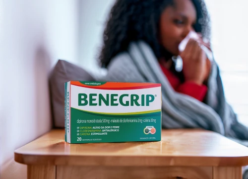 Benegrip em uma mesa na frente e mulher deitada na cama no fundo resfriada.